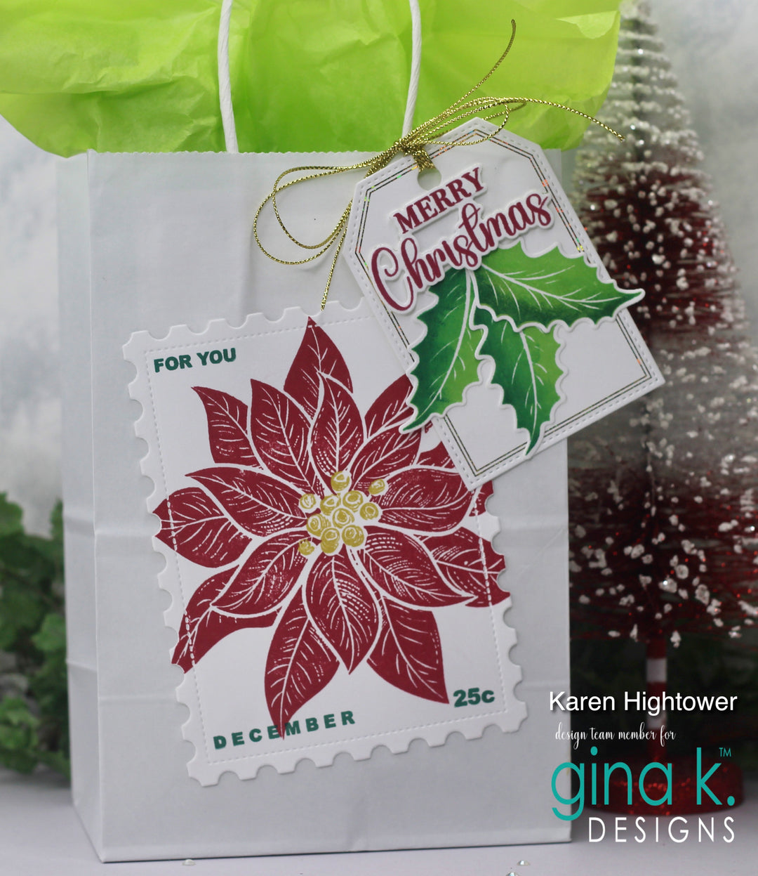 A white gift bag with a green tissue top and a gold ribbon, featuring a die-cut postage stamp design of a red poinsettia with a hanging 'Merry Christmas' tag. The photo is branded with 'gina k.