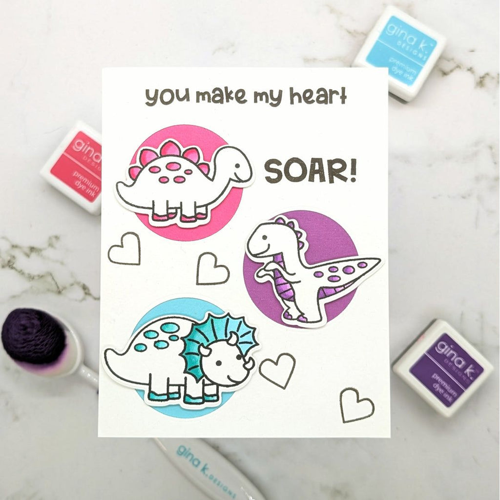 A white greeting card with a cute dinosaur design and the words 'you make my heart SOAR!' is displayed on a light marble surface, surrounded by Gina K. brand ink pads and a blending brush.