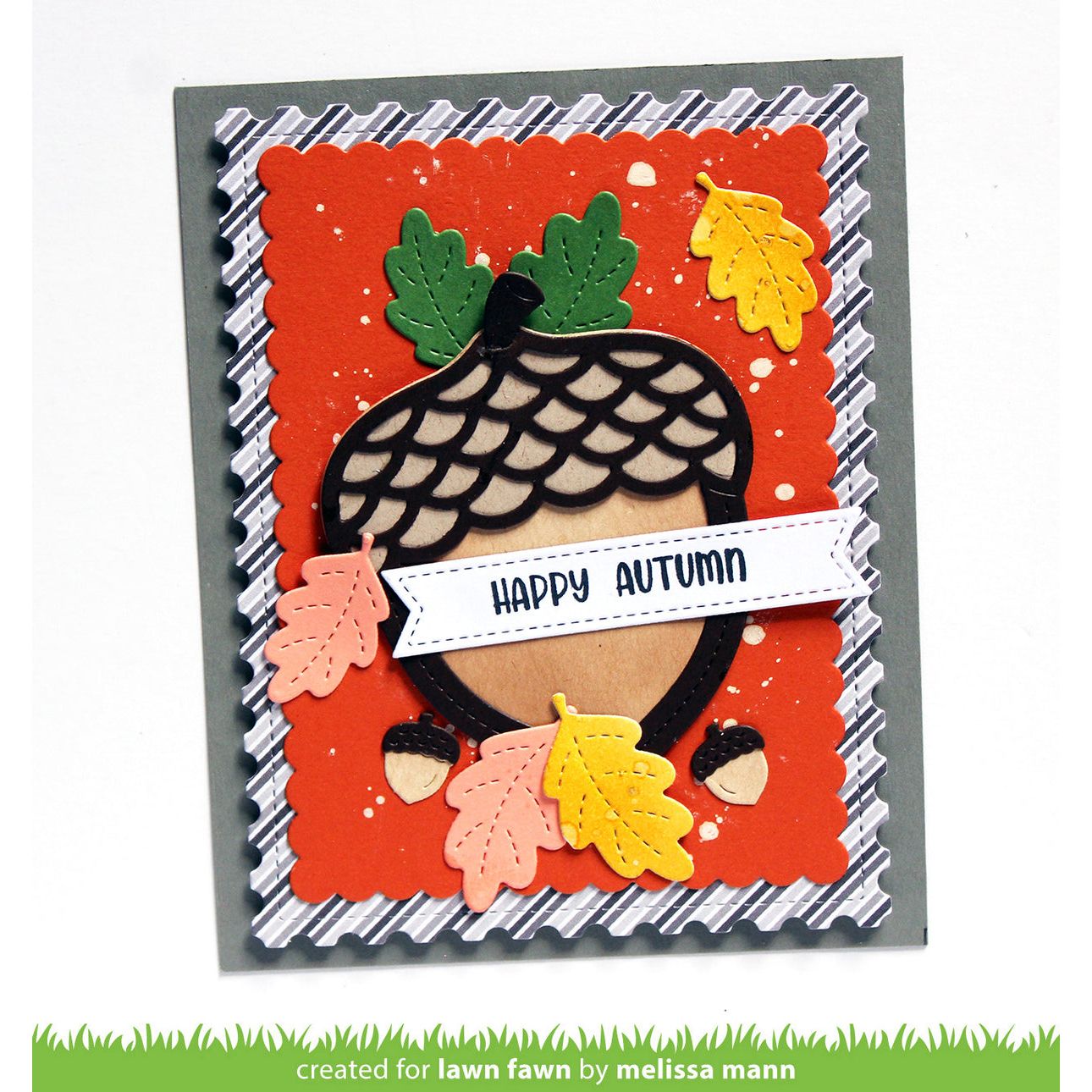 An autumn-themed layered card featuring a large acorn with green leaves, colorful paper leaves, and a white banner reading 'HAPPY AUTUMN' on an orange background, photographed on a white surface.