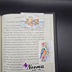 An open book page featuring a decorative printed bookmark or divider with colorful bird and floral illustrations, placed on a dark textured surface. A watermark reading 'Neema Design Team' is visible over the lower right area.