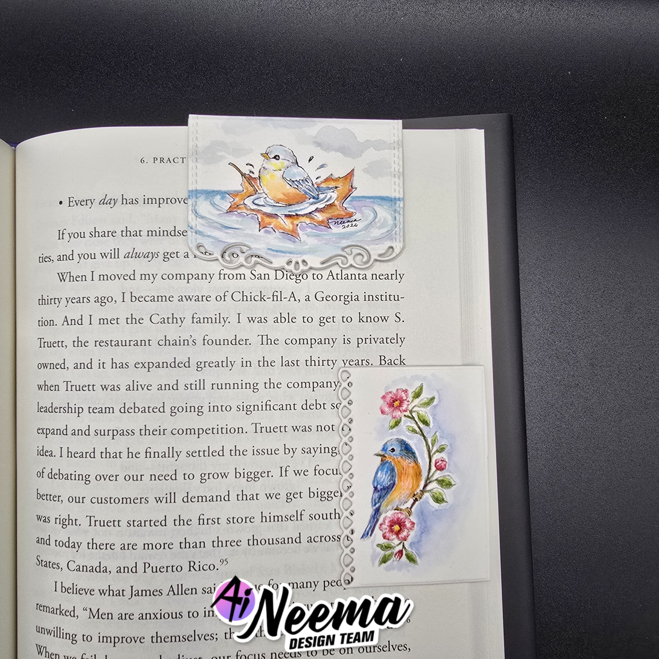 An open book page featuring a decorative printed bookmark or divider with colorful bird and floral illustrations, placed on a dark textured surface. A watermark reading 'Neema Design Team' is visible over the lower right area.