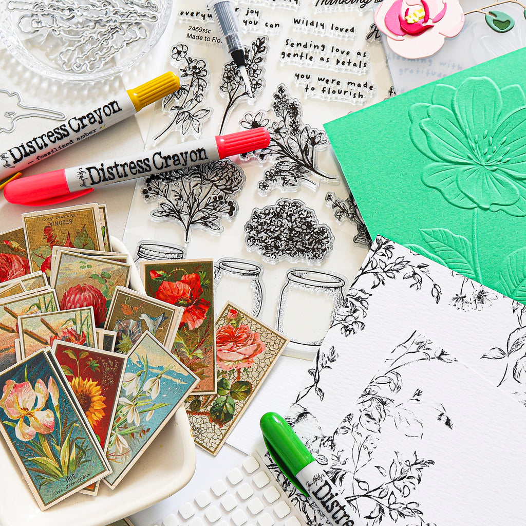 A flat-lay arrangement of distress crayons, clear stamp sheets with botanical and floral line art, vintage-style floral sticker cards, and a green embossed card on a white surface.
