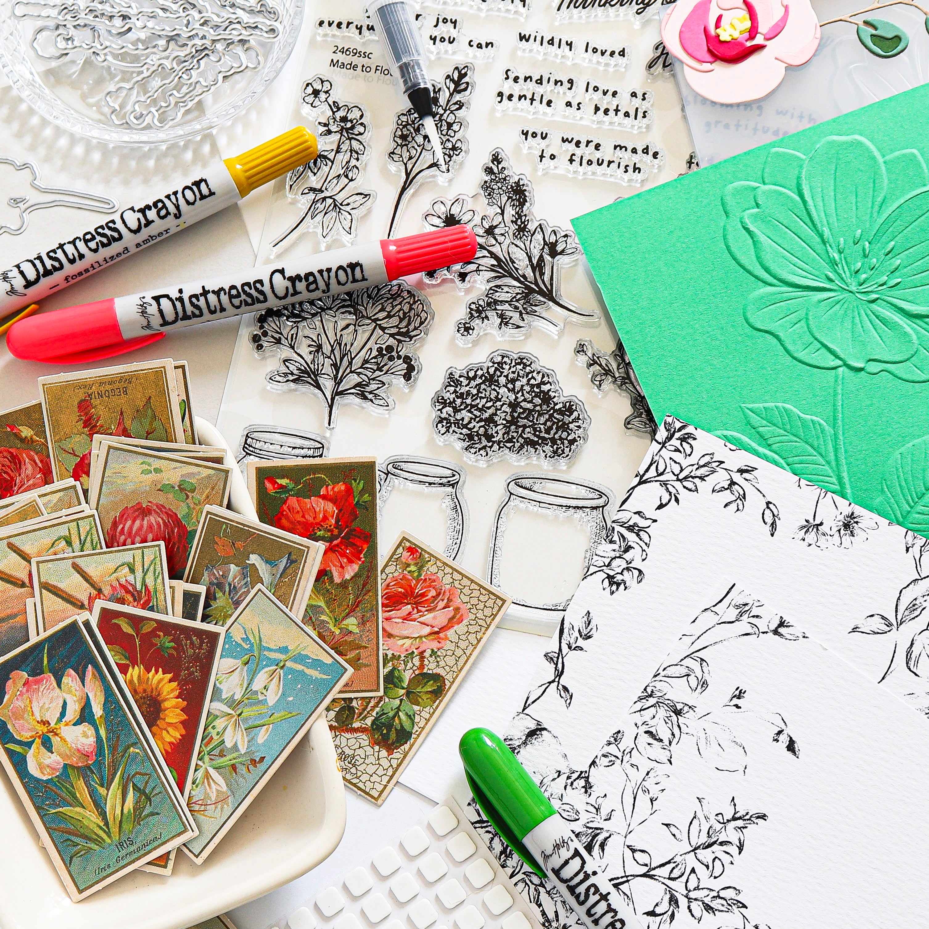 A flat-lay arrangement of distress crayons, clear stamp sheets with botanical and floral line art, vintage-style floral sticker cards, and a green embossed card on a white surface.