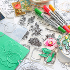A styled flat-lay of craft supplies on a white surface, including clear acrylic stamps with floral and jar illustrations, green and pink markers, metal cutters, a green embossed card, and decorative paper elements.