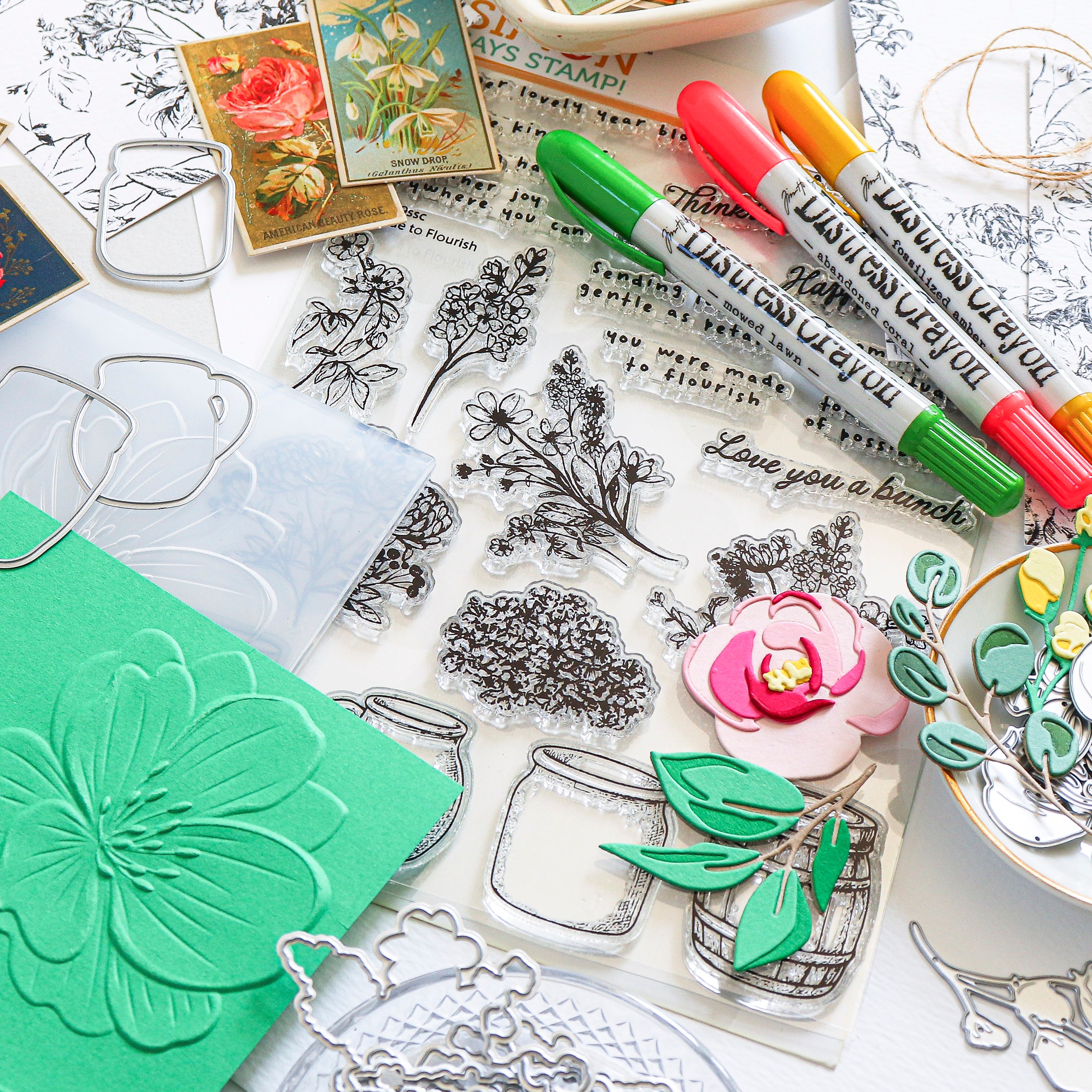 A styled flat-lay of craft supplies on a white surface, including clear acrylic stamps with floral and jar illustrations, green and pink markers, metal cutters, a green embossed card, and decorative paper elements.