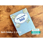 Card with 'World's Greatest Dad' text on a wooden surface, featuring Gina K Designs branding.