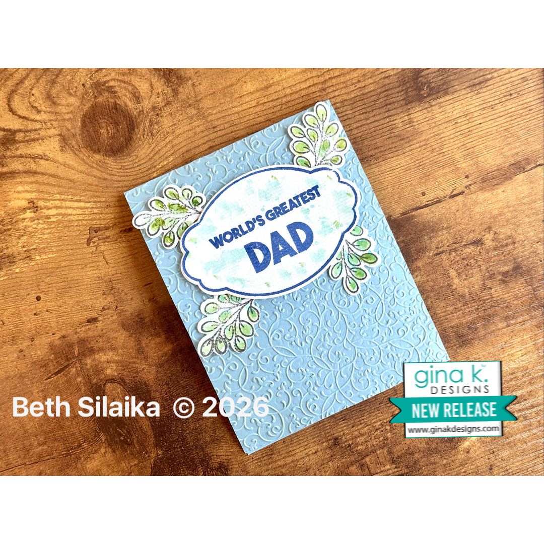 Card with 'World's Greatest Dad' text on a wooden surface, featuring Gina K Designs branding.