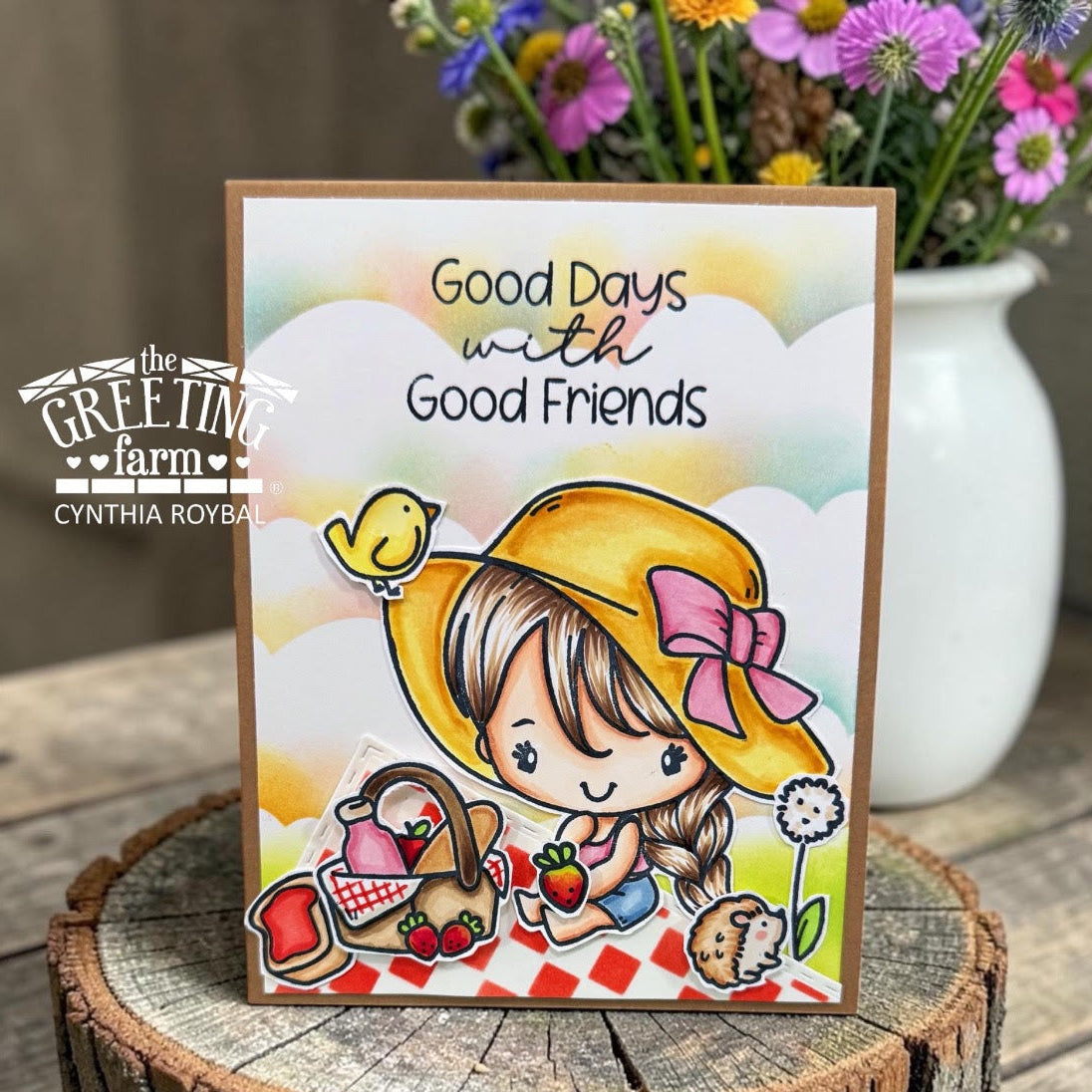 Card with a girl in a yellow hat and picnic basket, featuring 'Good Days with Good Friends' text, on a wooden surface with flowers in the background.