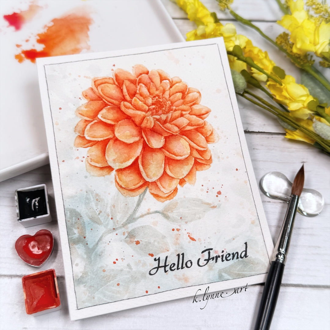 Card with a large orange flower and 'Hello Friend' text, surrounded by watercolor supplies on a wooden surface.