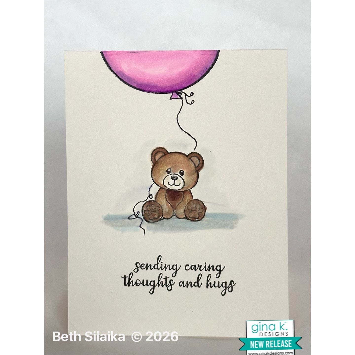 Card with a teddy bear holding a pink balloon and text 'sending caring thoughts and hugs' on a light background.