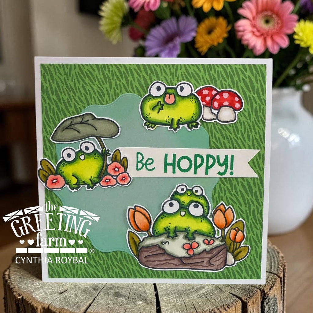 Card with cartoon frogs and flowers on a wooden block, featuring 'The Greeting Farm' branding.