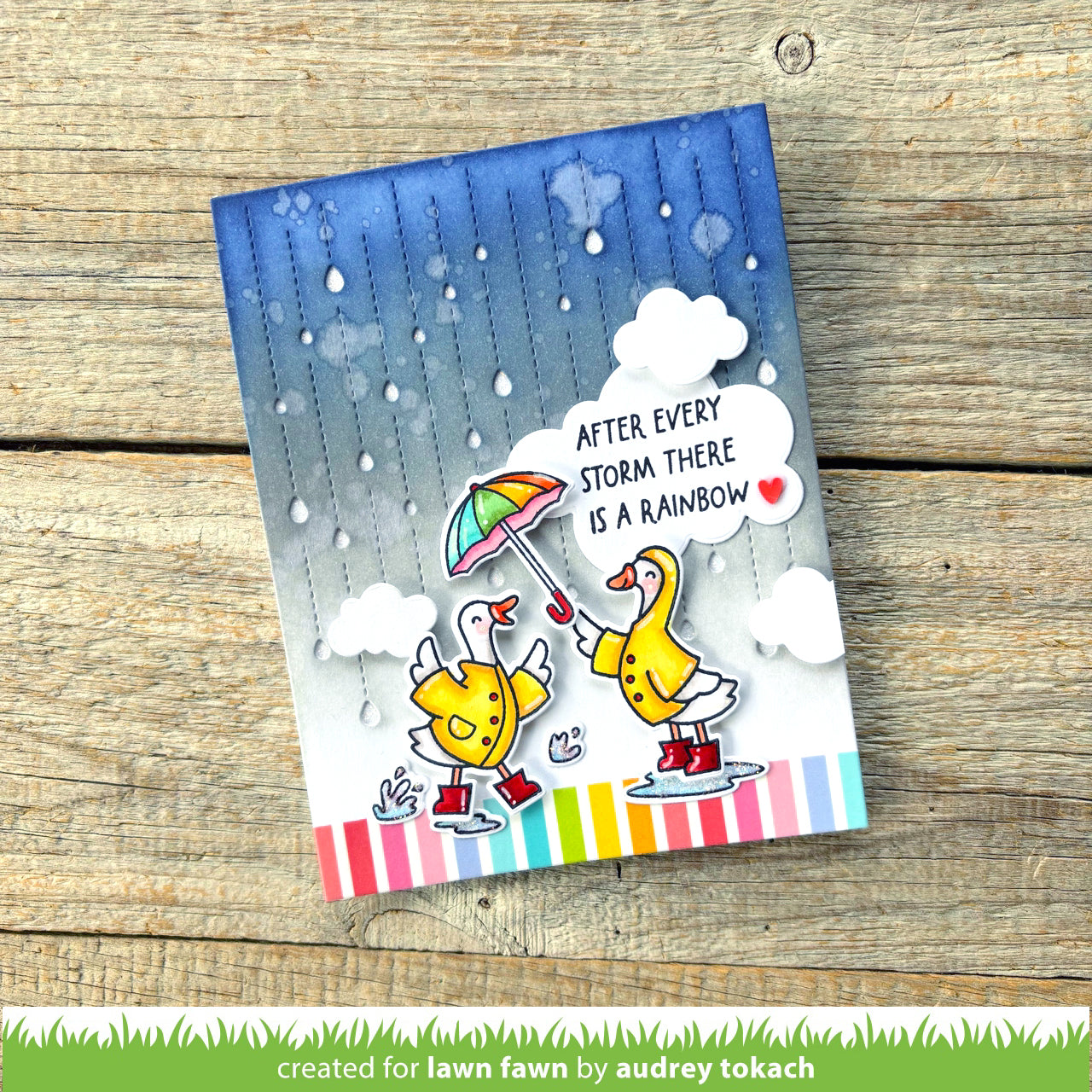 Card with cartoon geese under an umbrella and rainbow, text 'After every storm there is a rainbow', on a wooden surface.