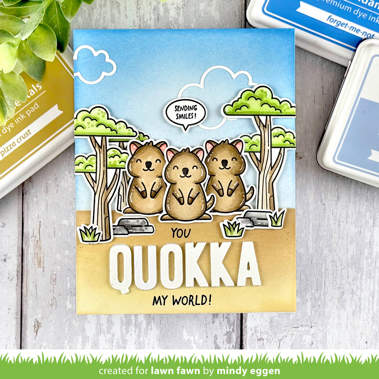 Card with cartoon quokkas and text 'You Quokka My World!' on a wooden surface with stamping supplies.