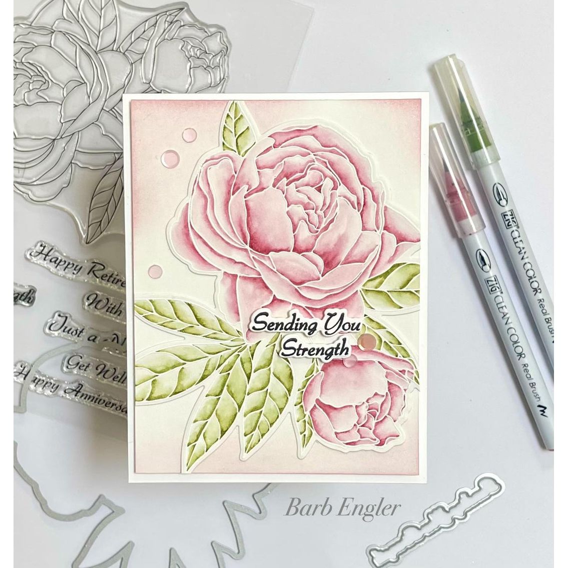 Card with pink peonies and 'Sending You Strength' text, surrounded by stamping tools on a white background.