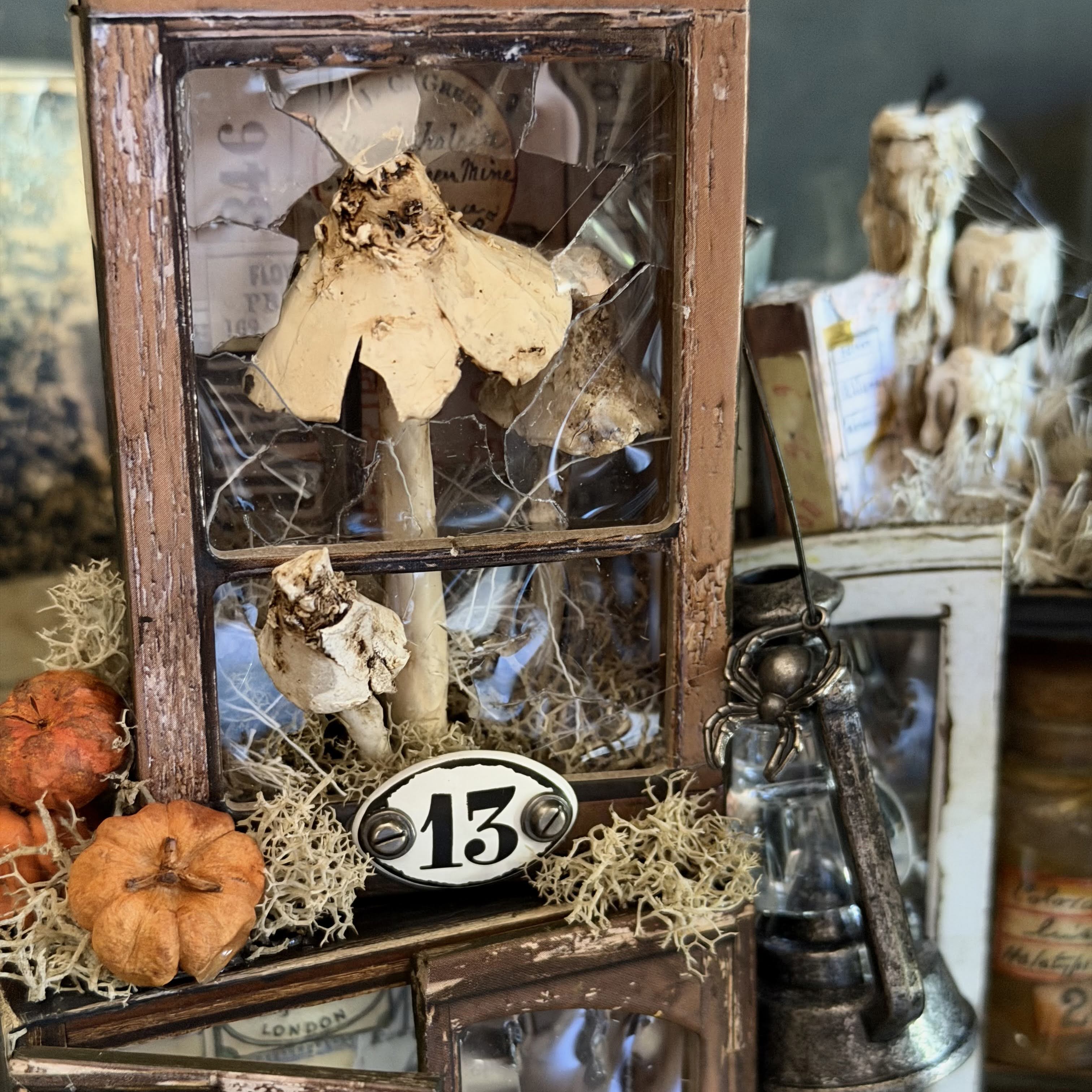 A rustic decorative display featuring a distressed wooden window frame with glass, filled with dried leaves, small orange pumpkins, beige moss, and a white oval sign reading “13.” A vintage lantern and additional autumn decor are visible in the softly blurred background.