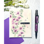Floral card with 'Thinking of You' message, purple pen, and purple envelope on textured white background