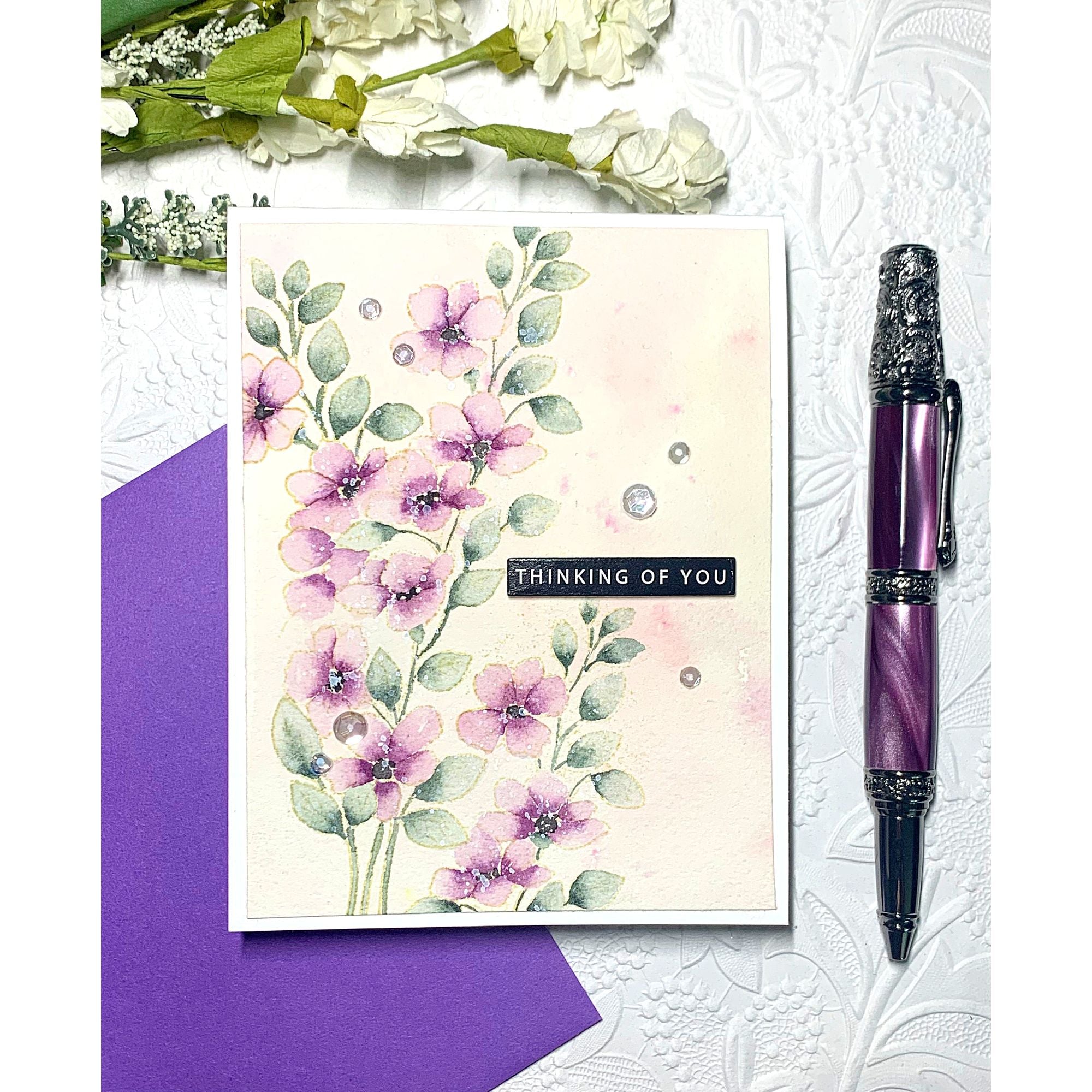 Floral card with 'Thinking of You' message, purple pen, and purple envelope on textured white background