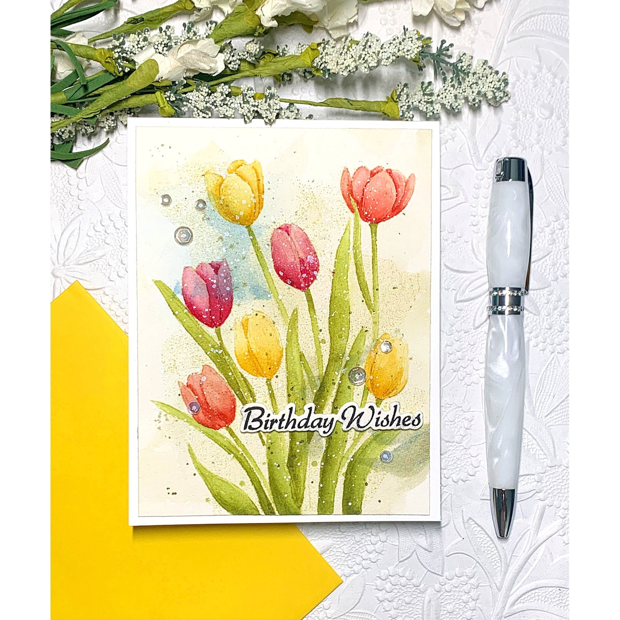 Greeting card with tulips and 'Birthday Wishes', white pen, and yellow envelope on textured surface.