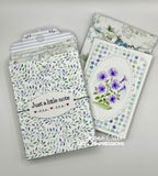 Handmade paper craft envelopes and a matching card featuring purple flowers, displayed on a light gray background. The card has a white base with a blue and purple floral oval design, and the envelopes feature a green leaf pattern, blue stripes, and a white label reading 'Just a little note'.