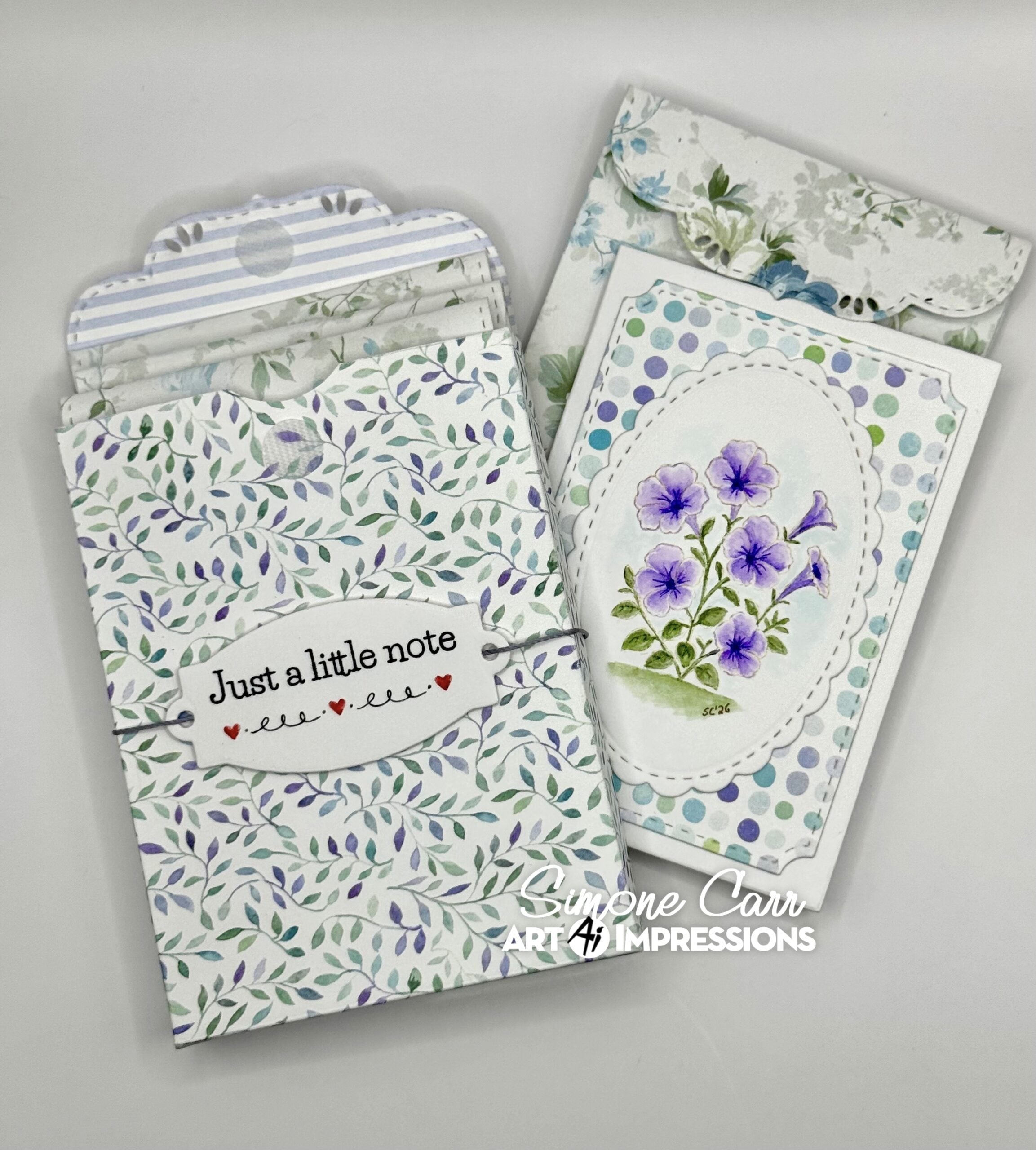 Handmade paper craft envelopes and a matching card featuring purple flowers, displayed on a light gray background. The card has a white base with a blue and purple floral oval design, and the envelopes feature a green leaf pattern, blue stripes, and a white label reading 'Just a little note'.