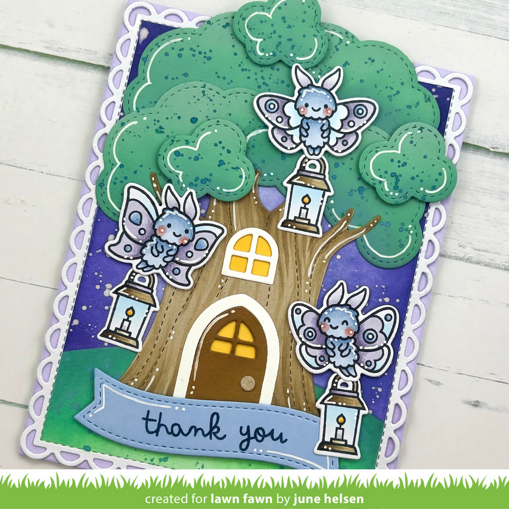 A handmade layered greeting card featuring a whimsical treehouse design with three cute blue-and-white fairy characters perched on lanterns. The card has a scalloped white border, a purple starry sky background, and a green grassy base.