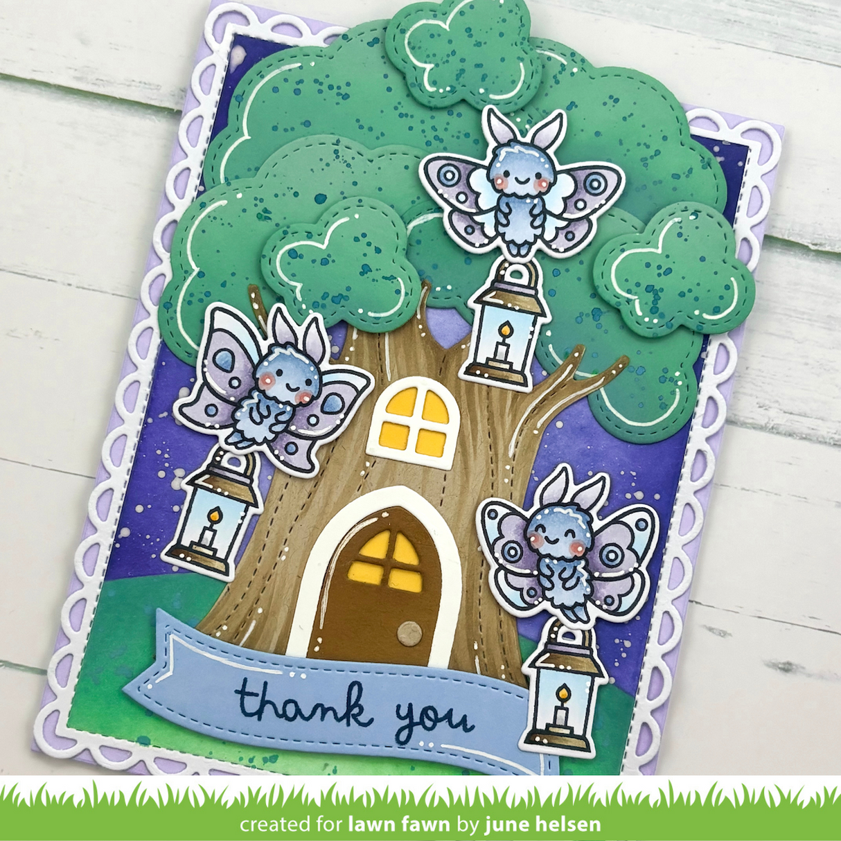 A handmade layered greeting card featuring a whimsical treehouse design with three cute blue-and-white fairy characters perched on lanterns. The card has a scalloped white border, a purple starry sky background, and a green grassy base.