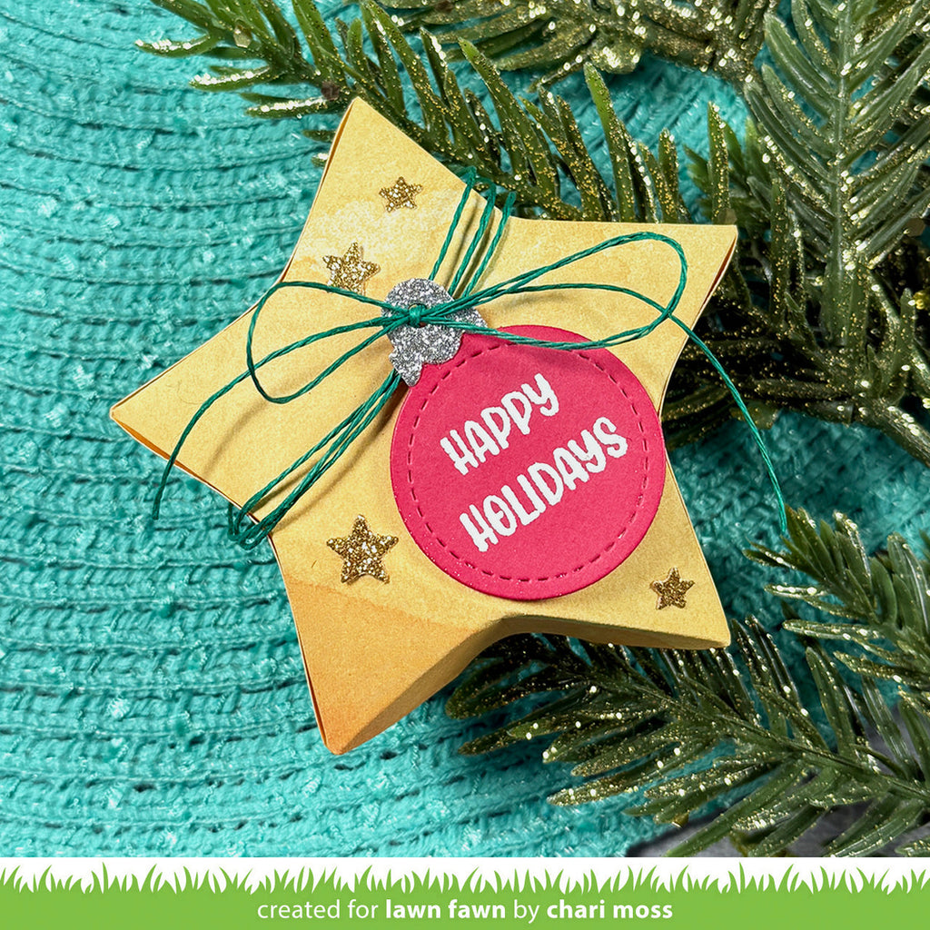 Star-shaped decorative item with 'Happy Holidays' tag on a textured teal background with greenery.