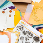 A styled flat-lay of craft supplies, including Simon Says Stamp! clear stamps with autumn-themed designs like a pumpkin truck and leaves, layered cardstock sheets in orange, yellow, teal, and purple, and paper tags on a neutral background.