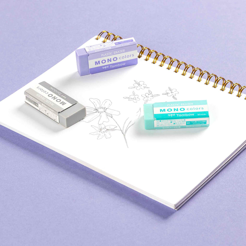 Three MONO colors erasers in purple, gray, and mint green are arranged on an open white spiral notebook with floral line art, set against a solid lavender background.