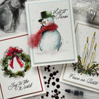 Three handmade greeting cards arranged on a glittery white surface with crafting props. The center card shows a watercolor snowman with a red scarf and the words “Let it Snow.