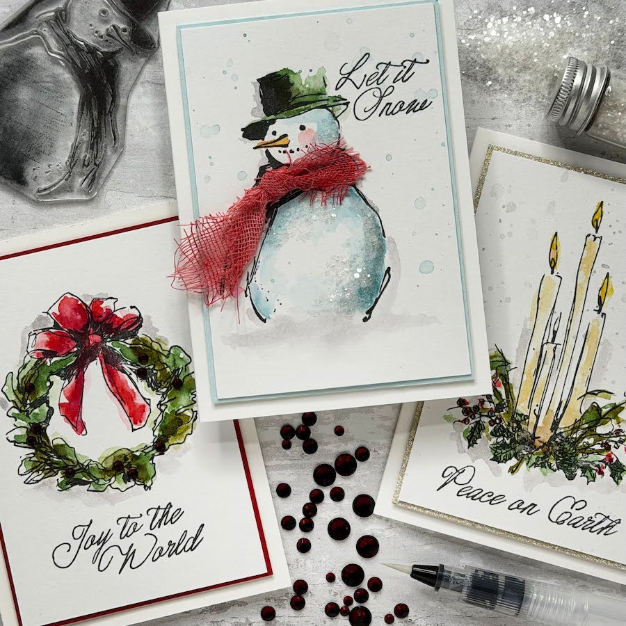 Three handmade greeting cards arranged on a glittery white surface with crafting props. The center card shows a watercolor snowman with a red scarf and the words “Let it Snow.