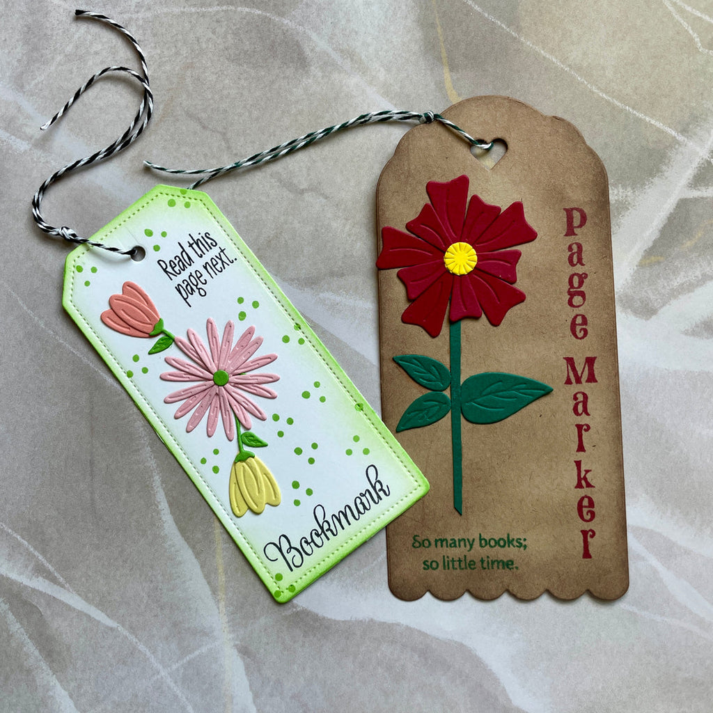 Two decorative bookmarks with printed text and colorful flower embellishments, photographed on a light beige fabric background. One bookmark is white with green dotted borders and pink and yellow flowers; the other is tan with a large red flower and the words 'Page Marker' and 'So many books; so little time.
