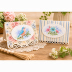 Two decorative handmade greeting cards displayed on a wooden surface with a lace doily and flowers. One card has a pink floral border and a central oval image of a blue bird.