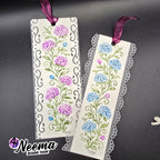 Two decorative paper bookmarks with floral designs, hanging from purple ribbons on a dark gray background. One bookmark has purple flowers and scrollwork, and the other has blue flowers and lace trim.