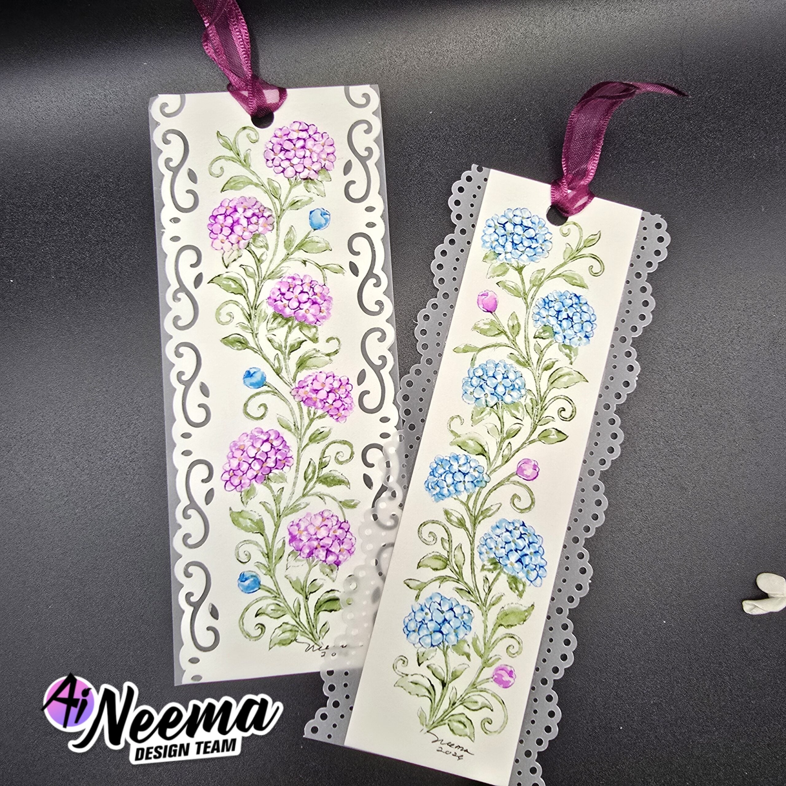 Two decorative paper bookmarks with floral designs, hanging from purple ribbons on a dark gray background. One bookmark has purple flowers and scrollwork, and the other has blue flowers and lace trim.