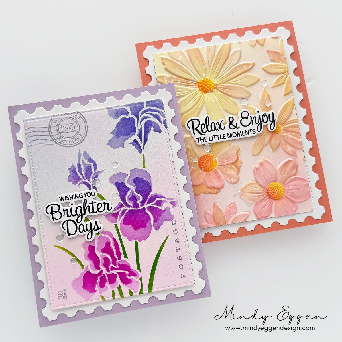 Two handmade postage-stamp-style greeting cards with floral designs, shown on a clean white background. One card has lavender and purple irises on a pale pink base, and the other has peach and yellow flowers on a coral base. | color-code:ALT01