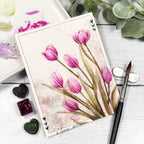 Watercolored pink tulips on a white card with art supplies and greenery.